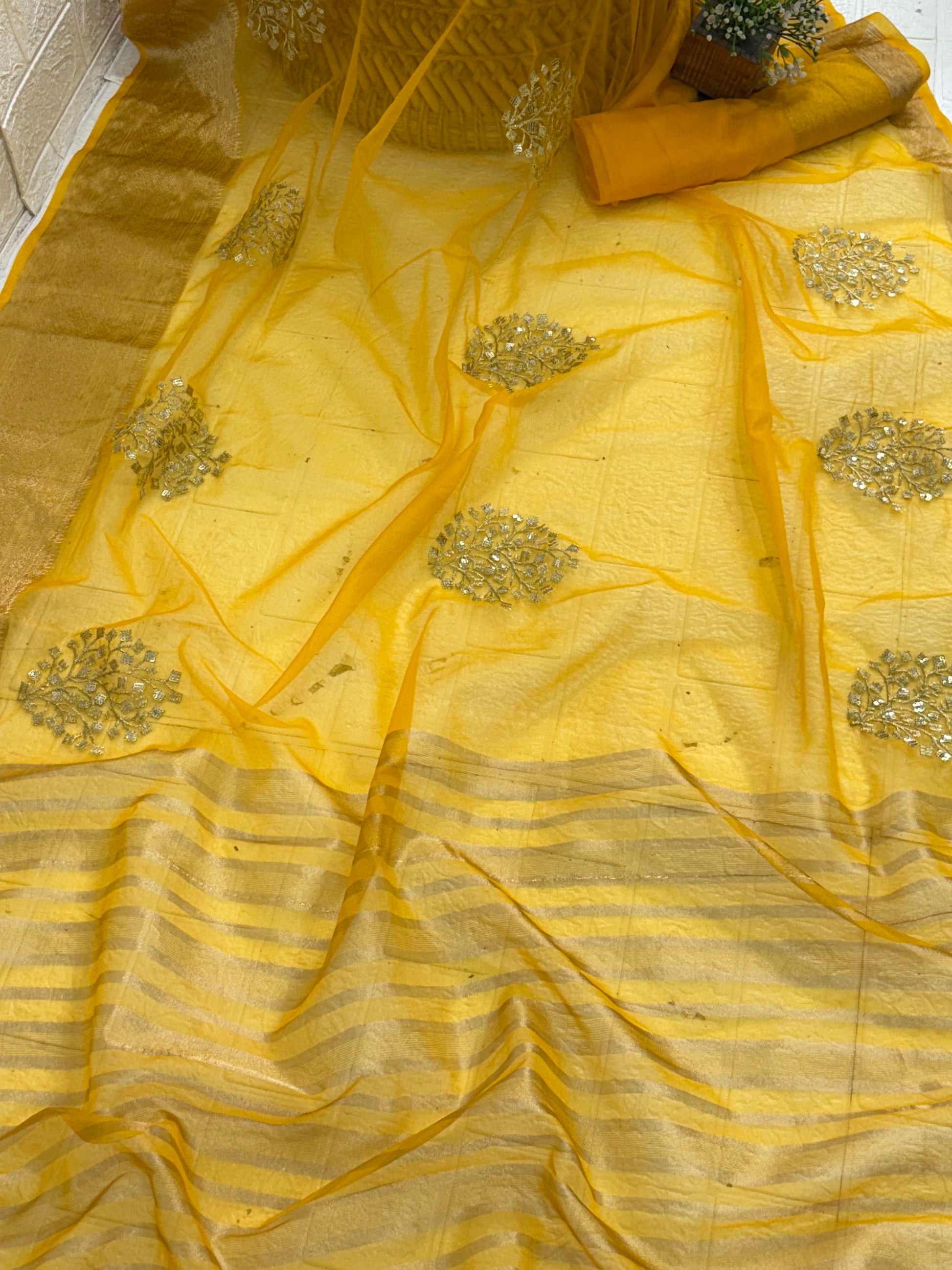 This is likely an Organza or fabric saree. The sheer, light-weight look suggests a fine fabric like Organza, or a similar silk blend, adorned with matching blouse golden zari lines on the pallu and intricate golden sequin or bead floral embroidery
