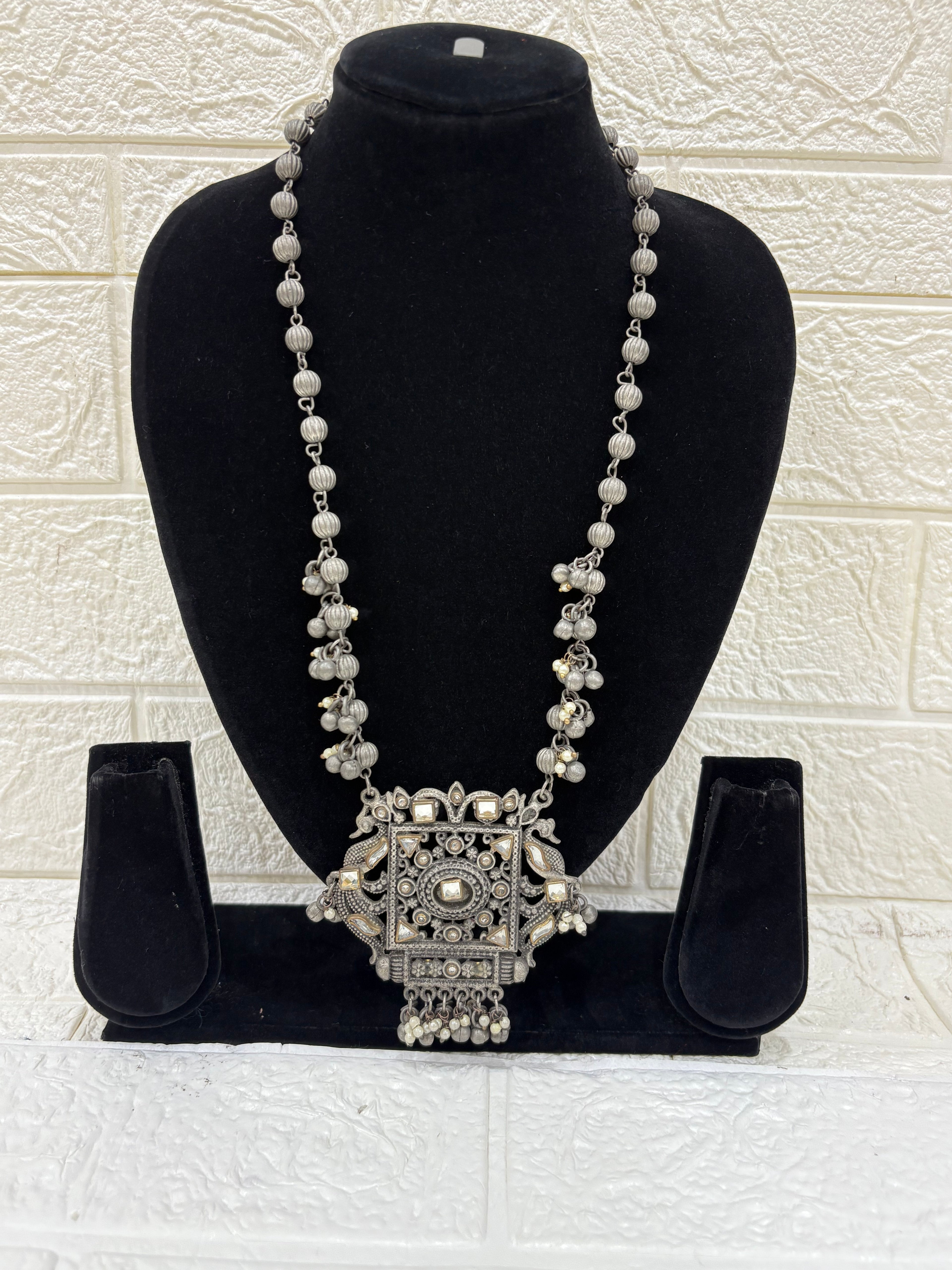 Name:
Antique Mirror Pendant Necklace Traditional oxidised silver necklace featuring a bold square mirror pendant with intricate detailing and dangling ghungroo accents for a statement festive look.