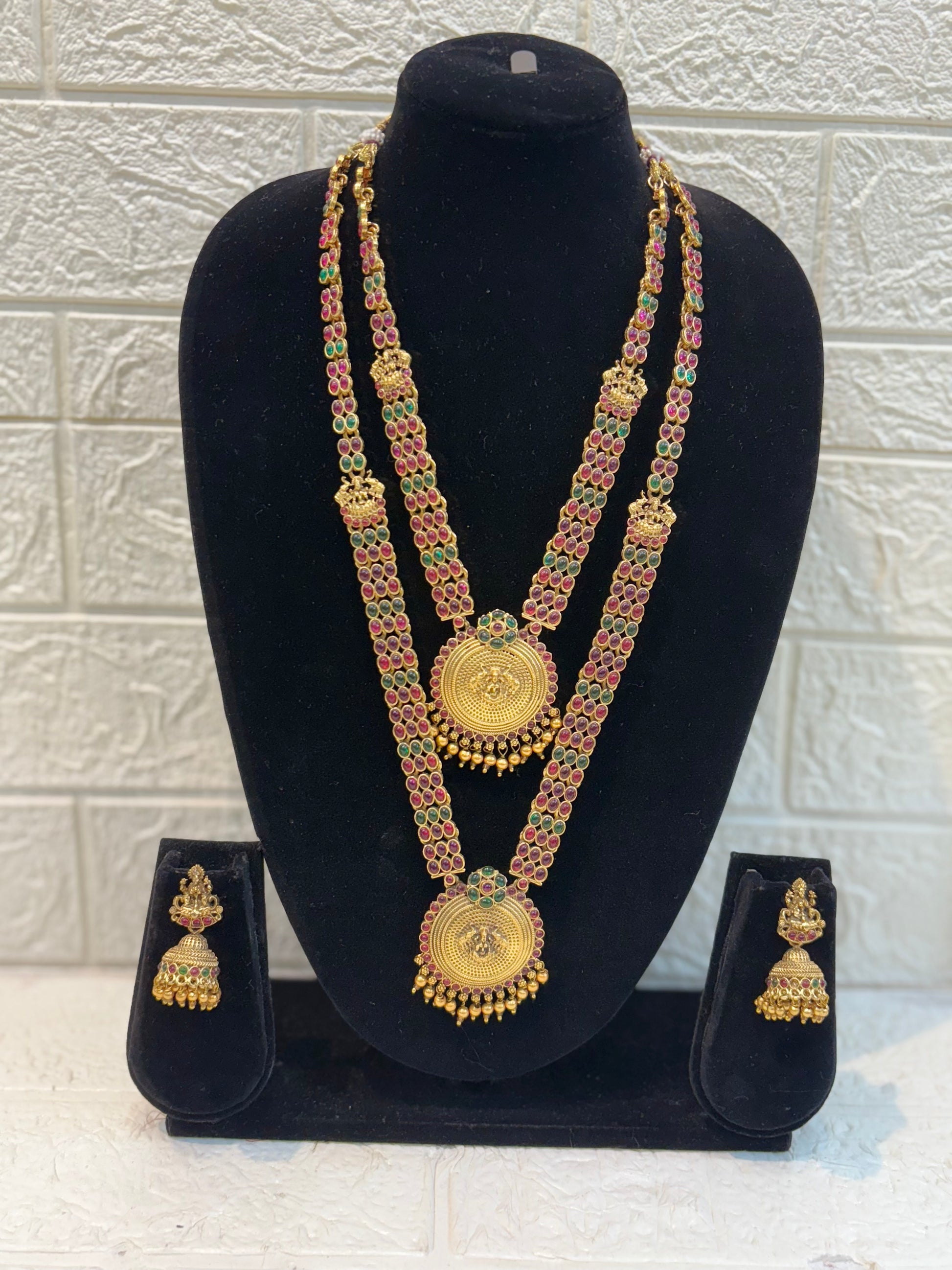 Elegant long haram featuring twin coin-style pendants with intricate embossing, ruby-green stone chain work, and traditional jhumka earrings—perfect for bridal and festive wear.
