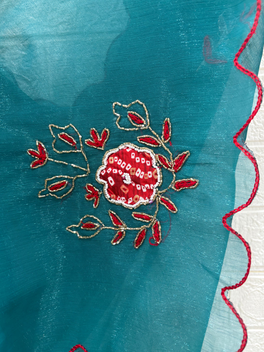 lightweight Teal Shimmer Organza with Embroidered material in a teal shade. It has a sheer, slightly shimmery texture with delicate embroidered floral motifs in red and gold scattered across the fabric. The border is finished with a fine red edg