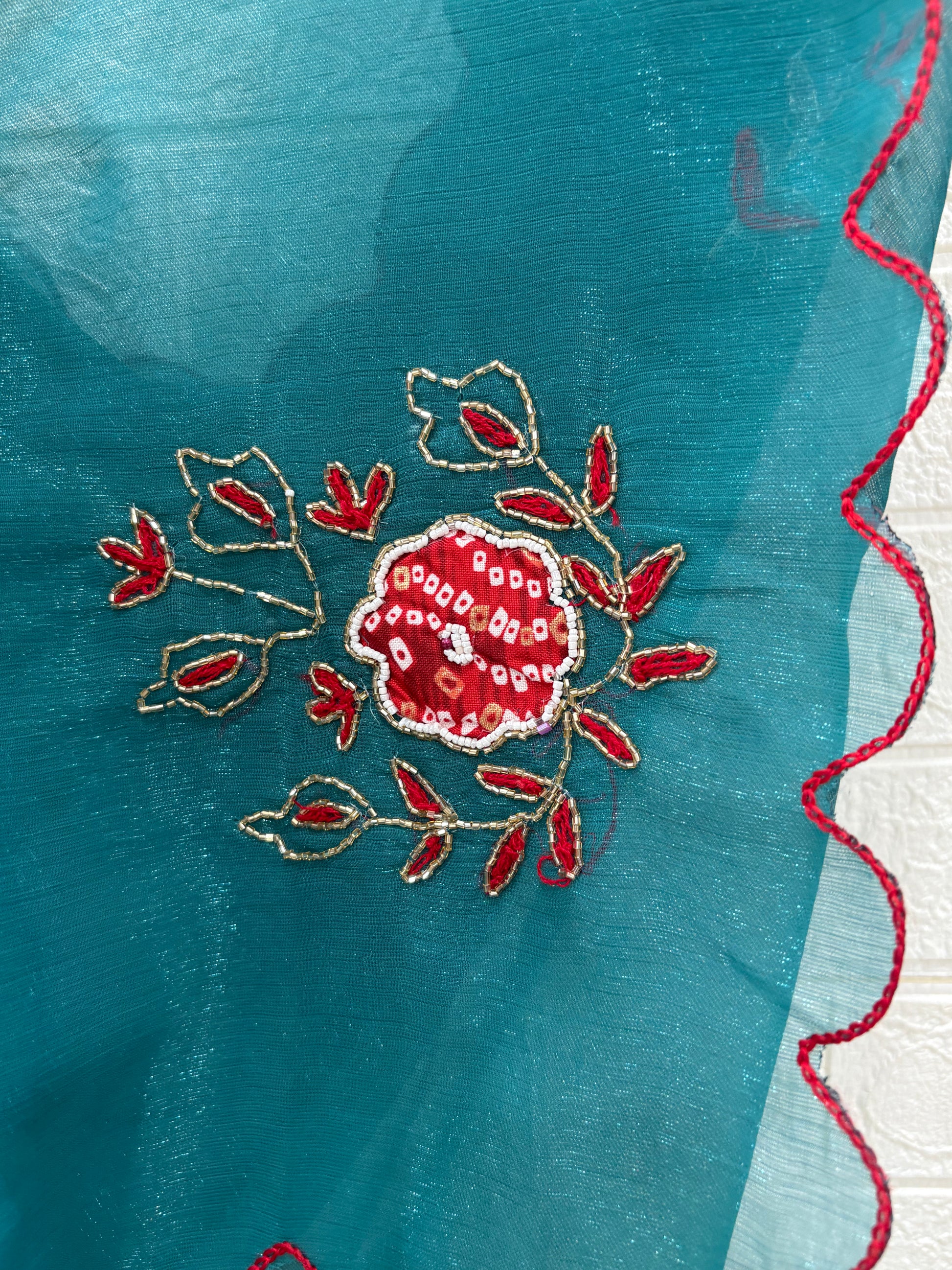 lightweight Teal Shimmer Organza with Embroidered material in a teal shade. It has a sheer, slightly shimmery texture with delicate embroidered floral motifs in red and gold scattered across the fabric. The border is finished with a fine red edg