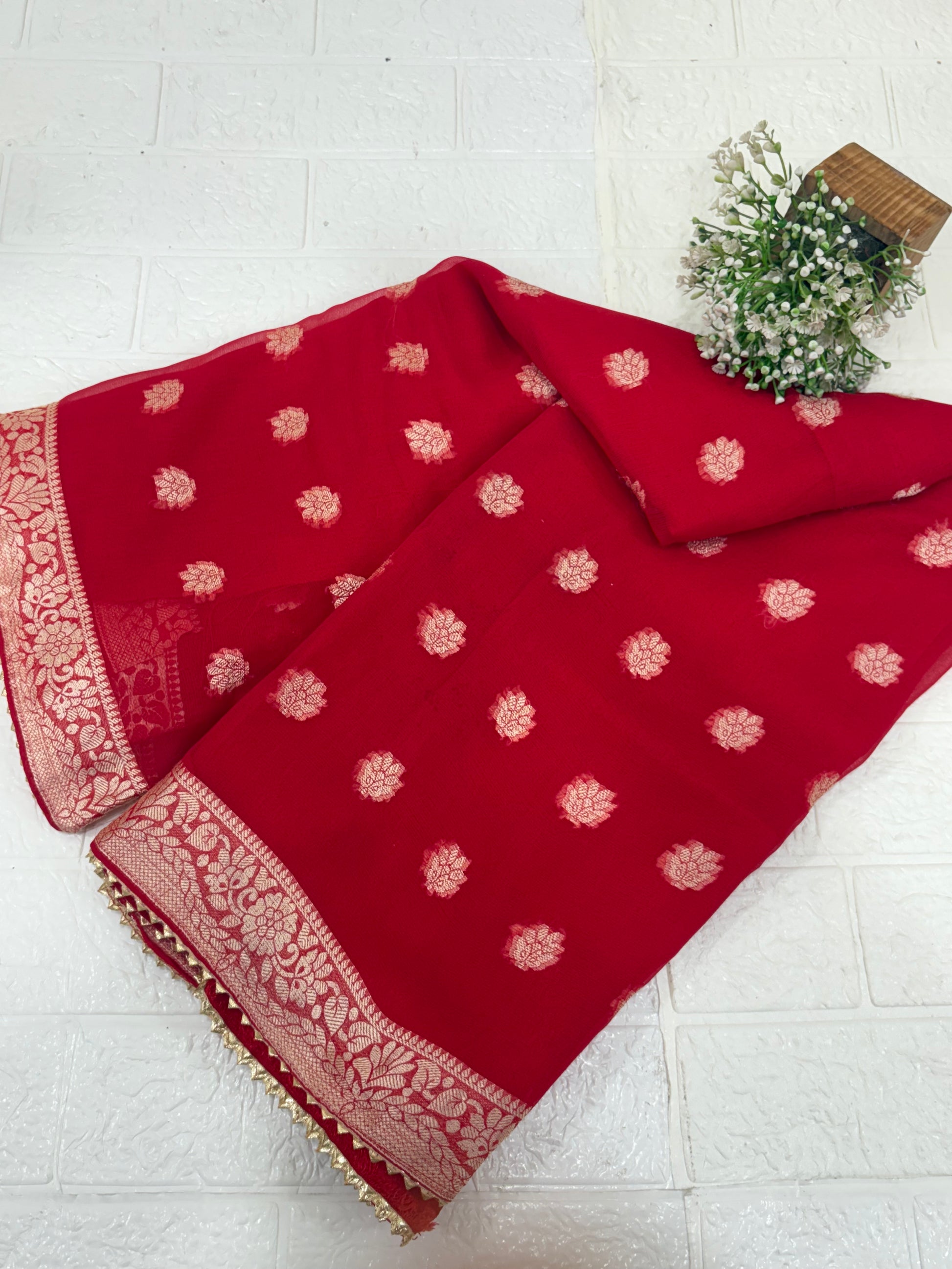 This fabric is a red Georgette with all-over gold zari floral buti embroidery and a rich woven border, paired with a matching plain blouse perfect for festival