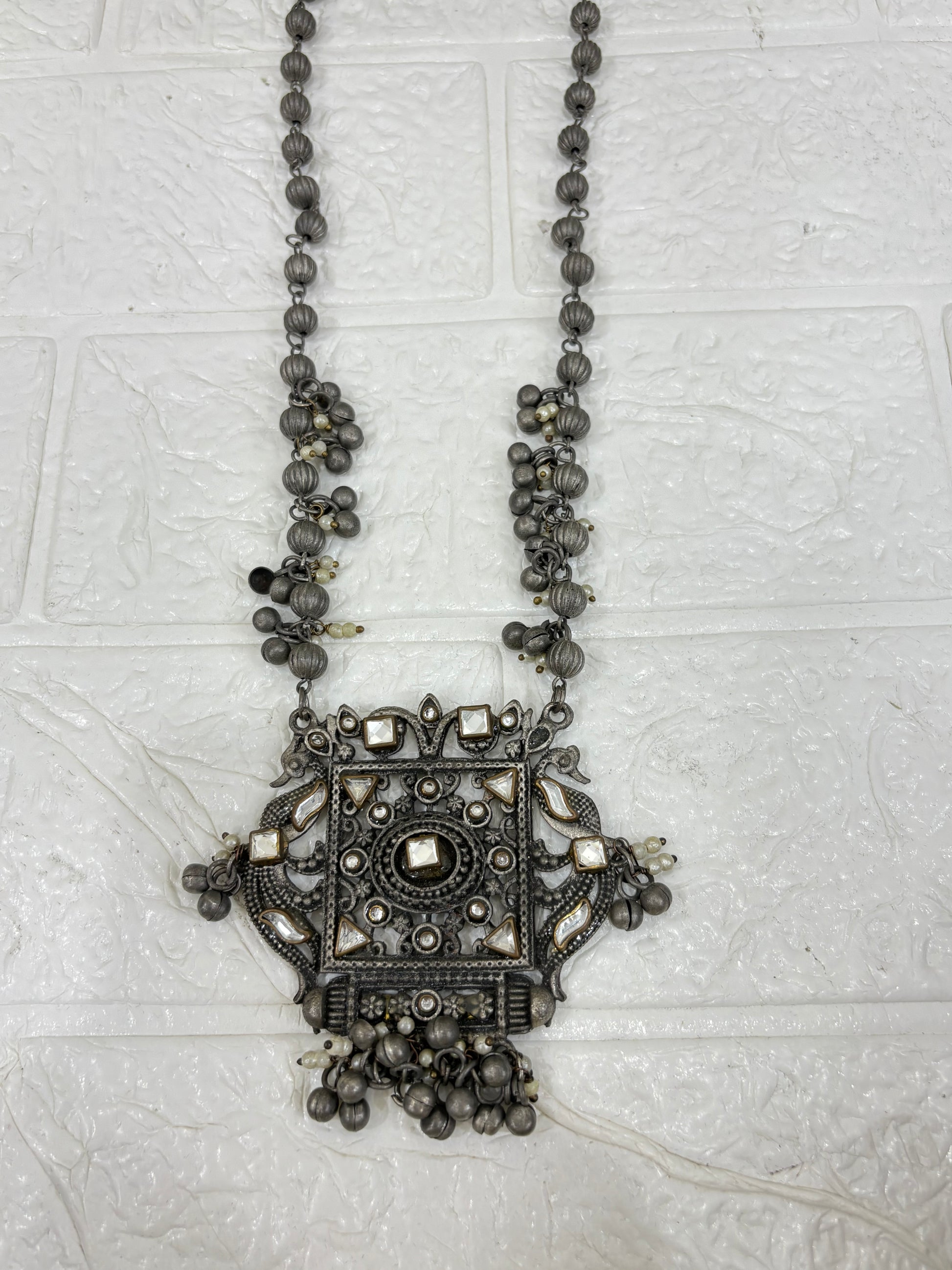 Name:
Antique Mirror Pendant Necklace Traditional oxidised silver necklace featuring a bold square mirror pendant with intricate detailing and dangling ghungroo accents for a statement festive look.