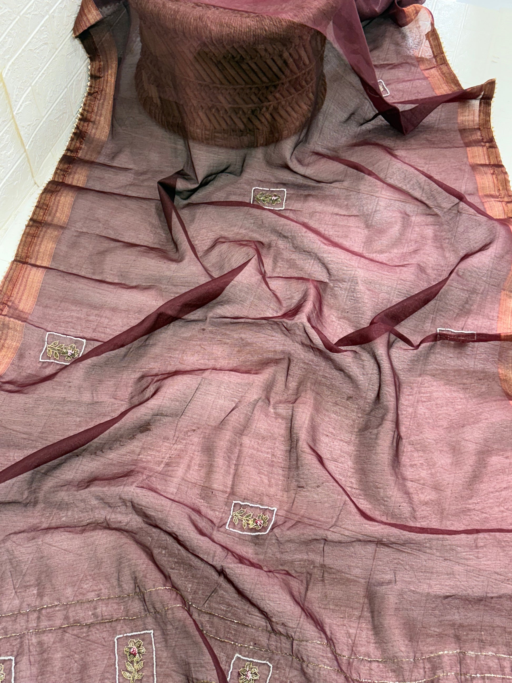 Name: Chanderi or Cotton Silk Saree Fabric The fine texture, sheen, and way the material is folded suggest a Chanderi or Cotton Silk Blend. The work is Zari Woven Stripes with Aari/Thread Embroidery motifs.