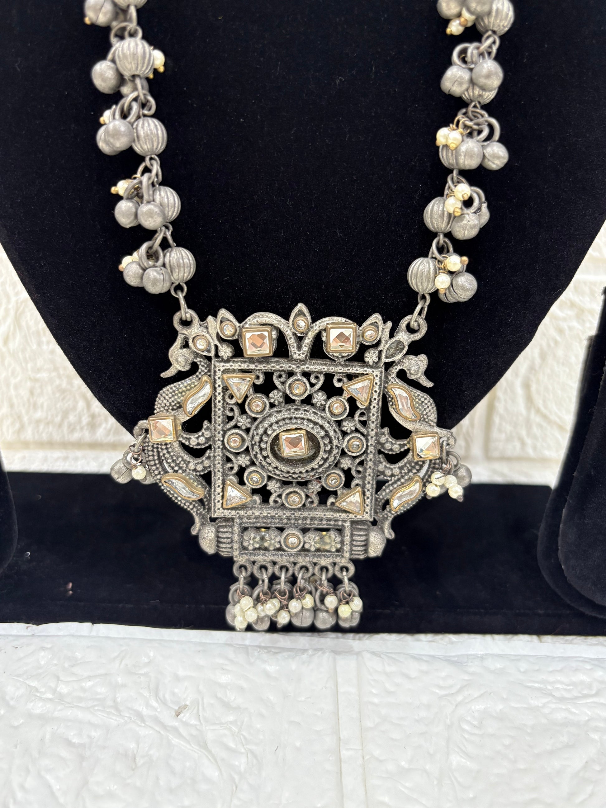 Name:
Antique Mirror Pendant Necklace Traditional oxidised silver necklace featuring a bold square mirror pendant with intricate detailing and dangling ghungroo accents for a statement festive look.