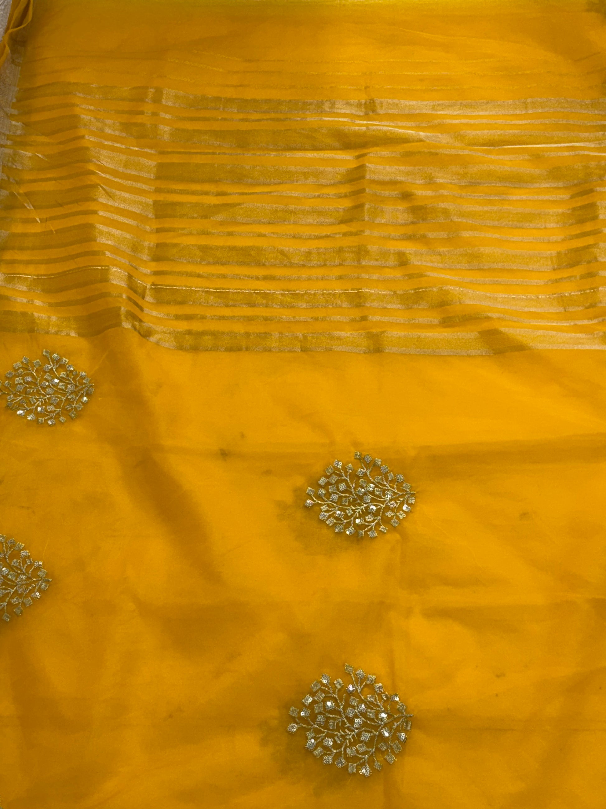 This is likely an Organza or fabric saree. The sheer, light-weight look suggests a fine fabric like Organza, or a similar silk blend, adorned with matching blouse golden zari lines on the pallu and intricate golden sequin or bead floral embroidery