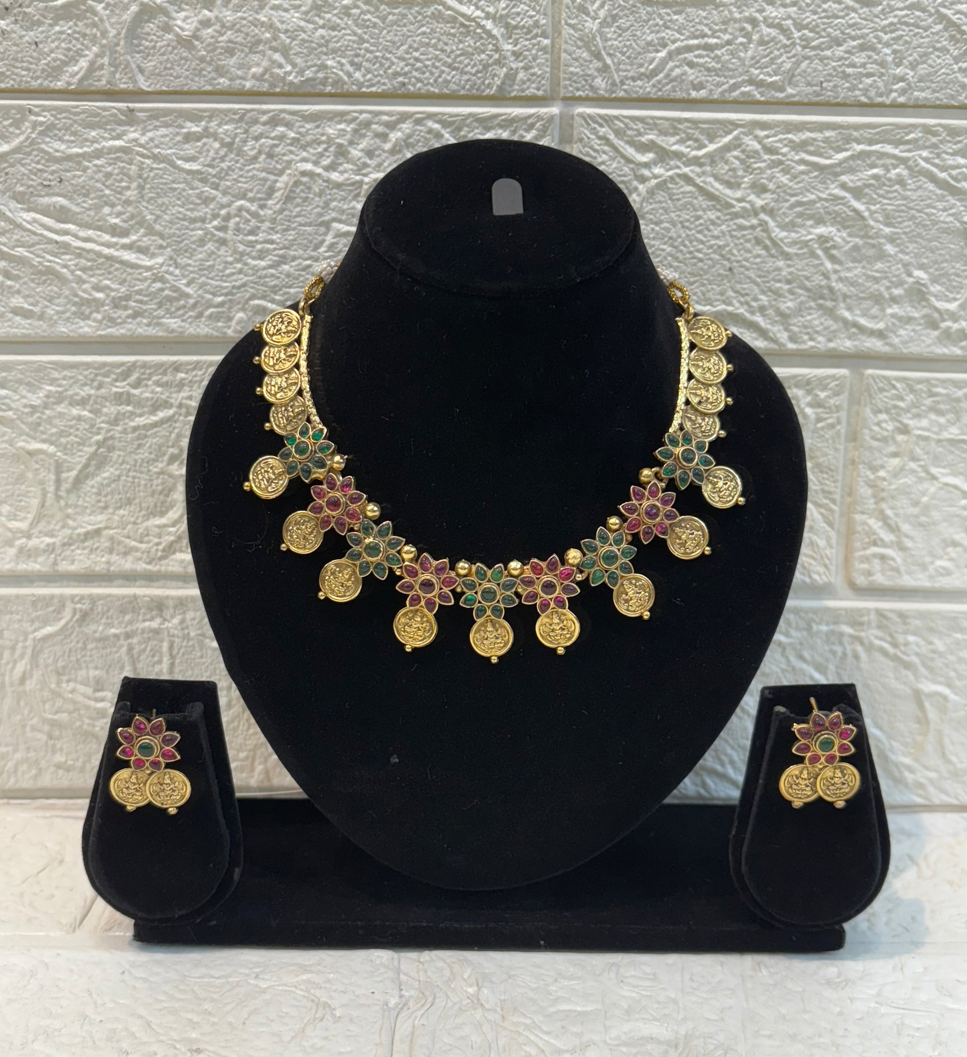 Traditional antique-finish necklace adorned with coin motifs, ruby-green floral stone work, and matching earrings—perfect for festive and wedding occasions.