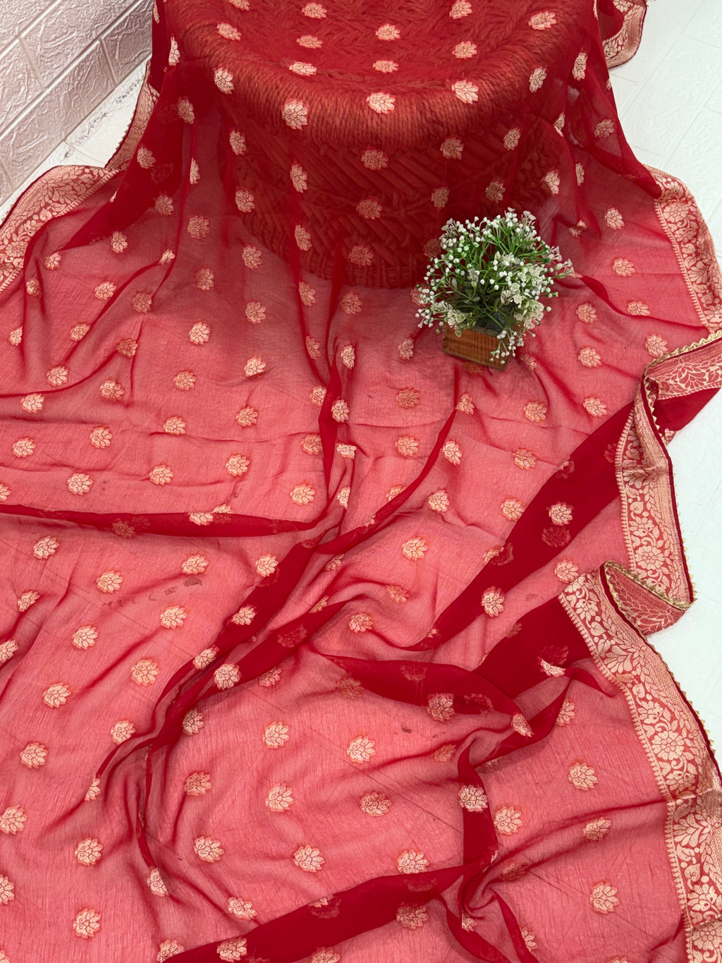 This fabric is a red Georgette with all-over gold zari floral buti embroidery and a rich woven border, paired with a matching plain blouse perfect for festival