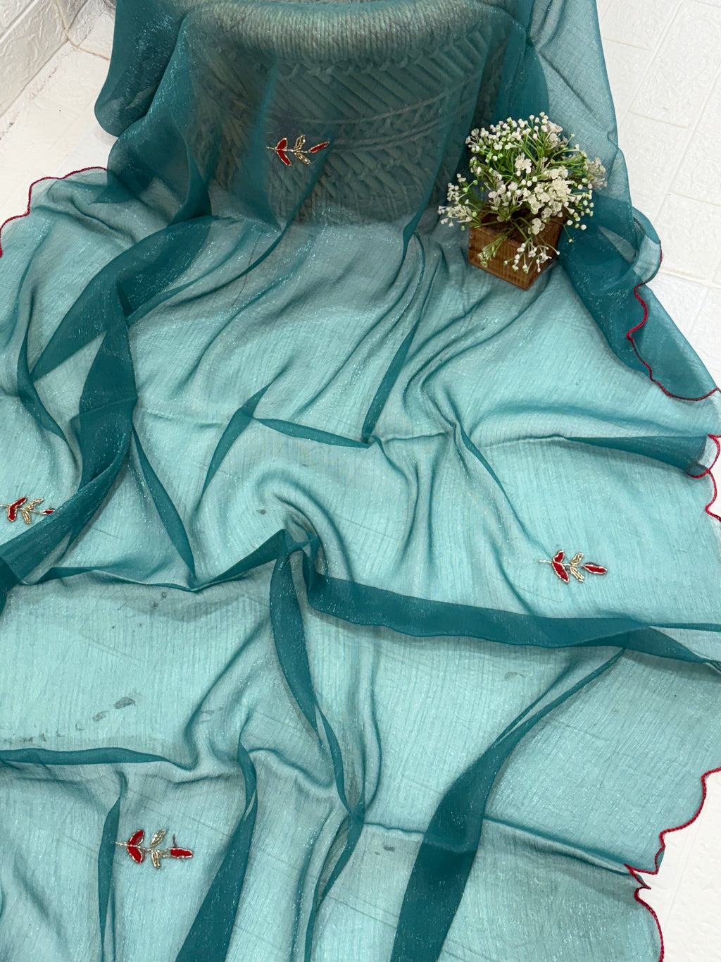 lightweight Teal Shimmer Organza with Embroidered material in a teal shade. It has a sheer, slightly shimmery texture with delicate embroidered floral motifs in red and gold scattered across the fabric. The border is finished with a fine red edg