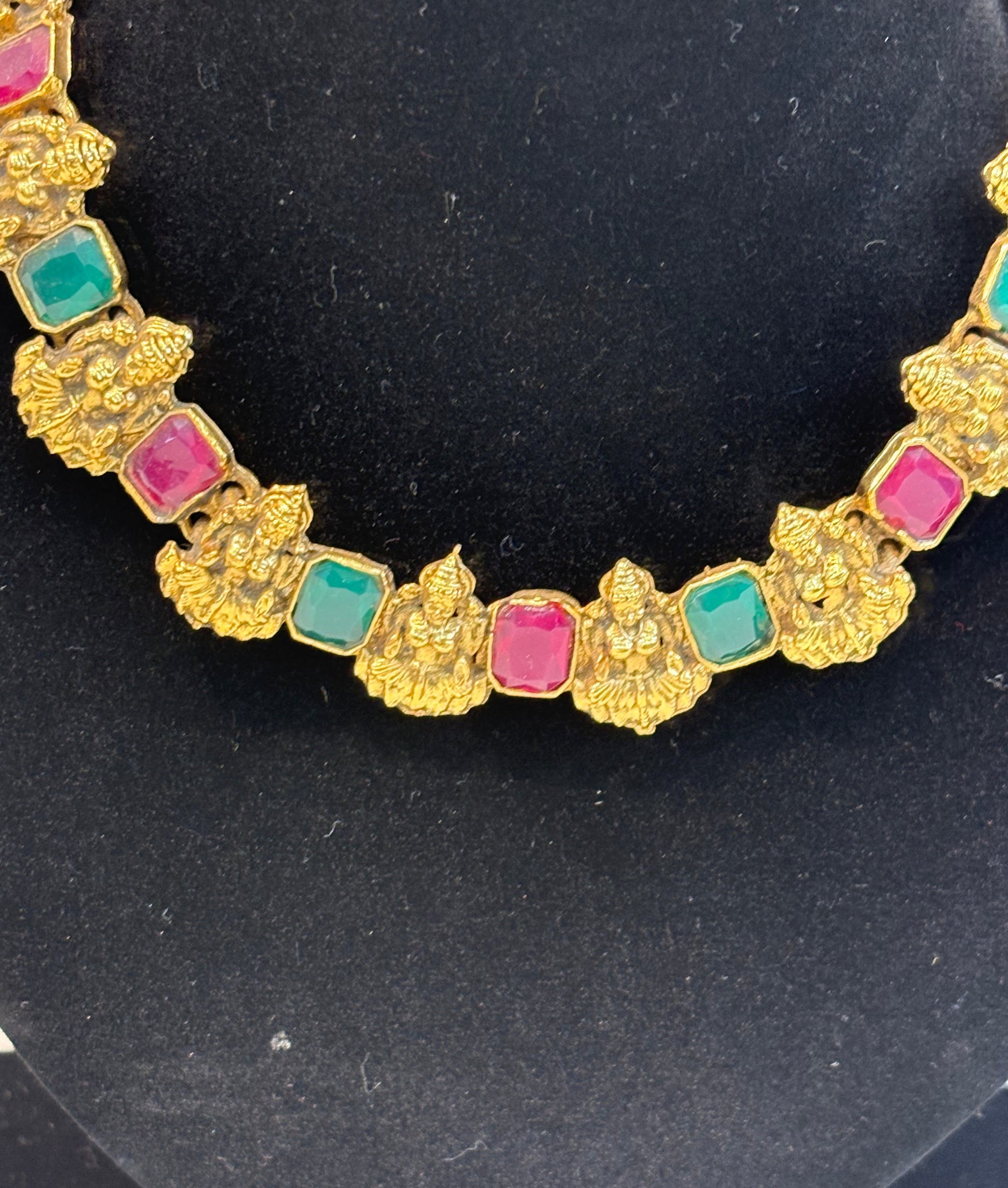 Classic temple-style necklace featuring Goddess Lakshmi motifs with ruby and green stone settings in an antique gold finish, paired with matching earrings—ideal for festive and bridal wear.