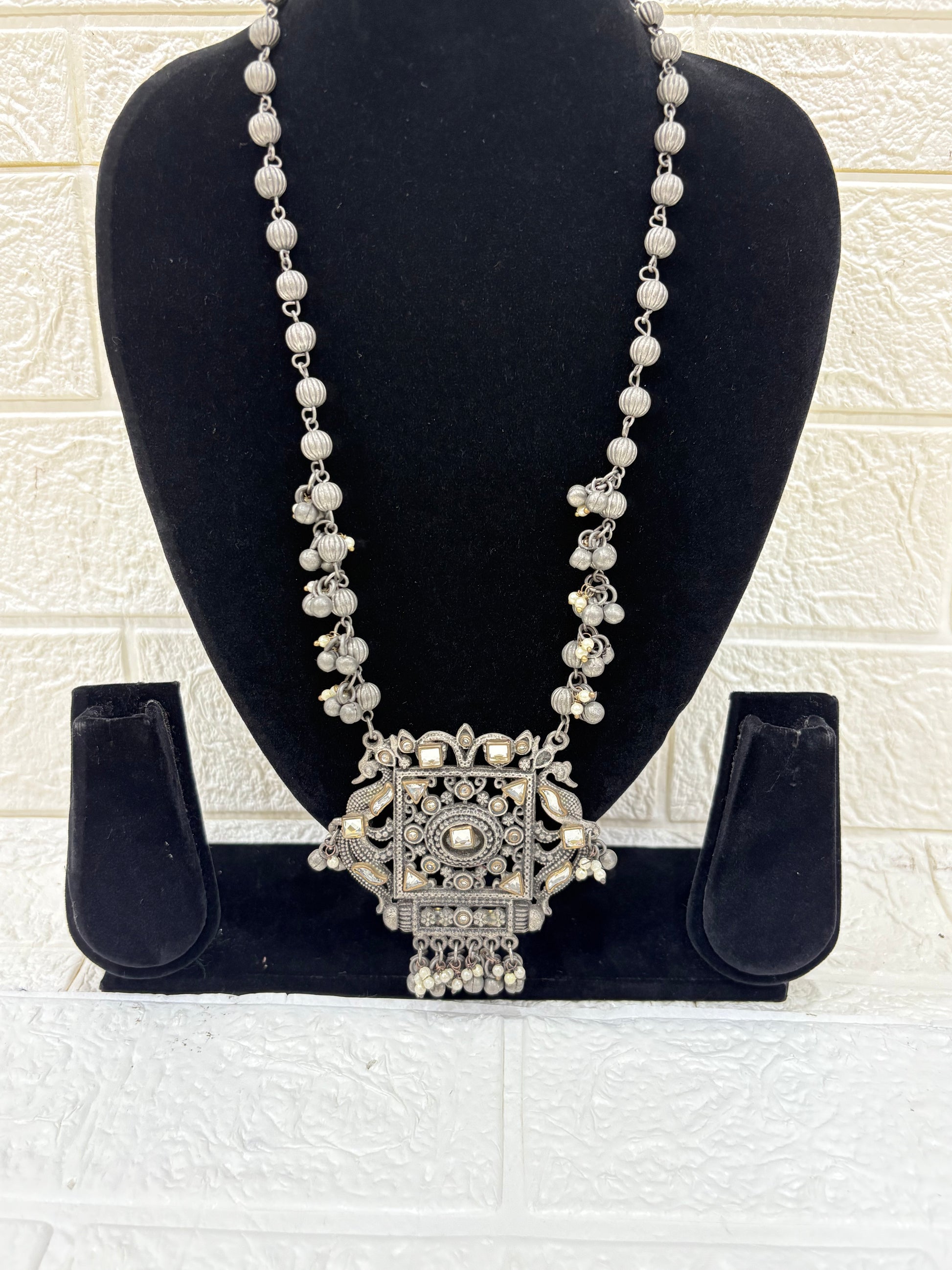 Name:
Antique Mirror Pendant Necklace Traditional oxidised silver necklace featuring a bold square mirror pendant with intricate detailing and dangling ghungroo accents for a statement festive look.