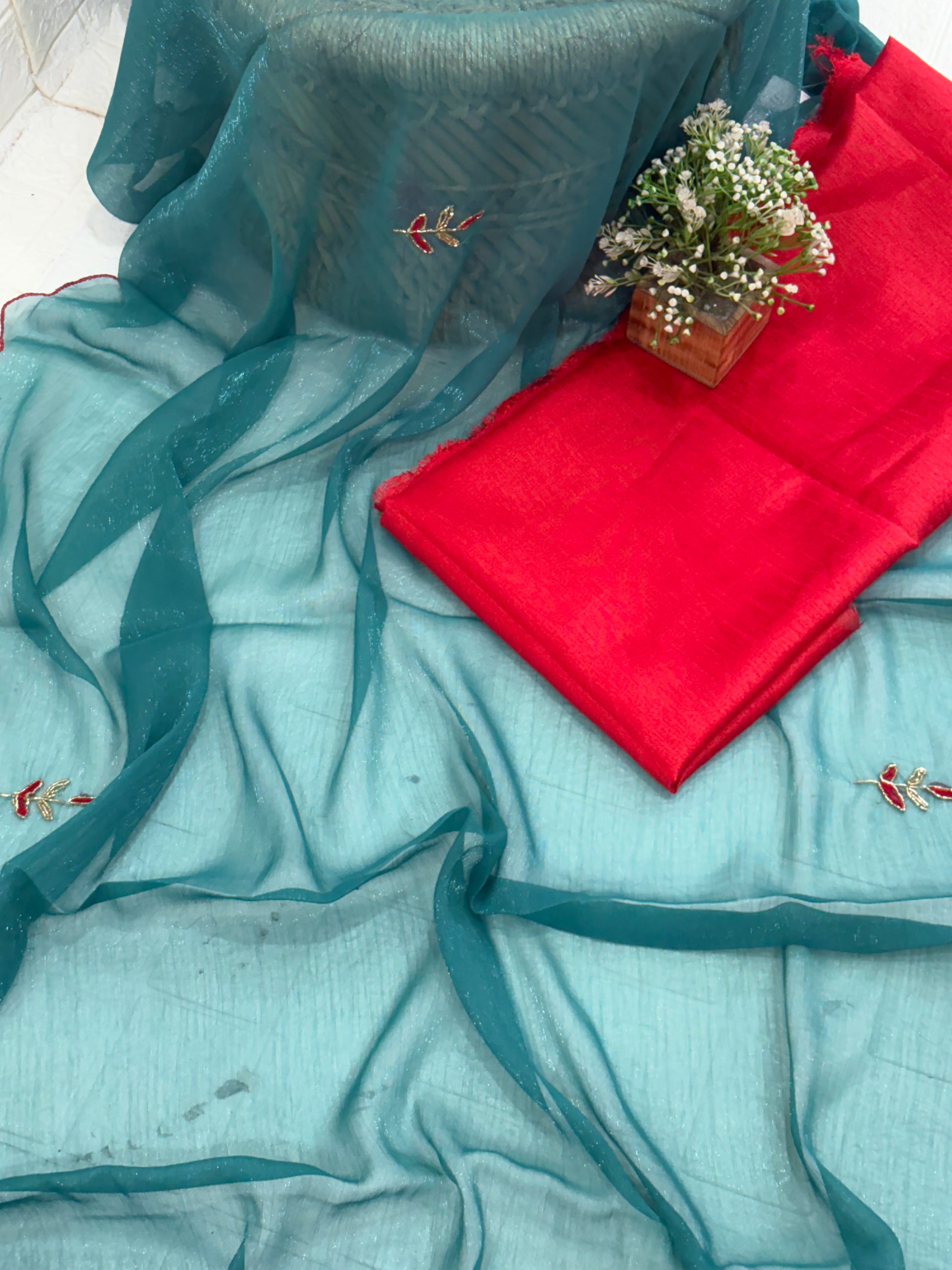 lightweight Teal Shimmer Organza with Embroidered material in a teal shade. It has a sheer, slightly shimmery texture with delicate embroidered floral motifs in red and gold scattered across the fabric. The border is finished with a fine red edg