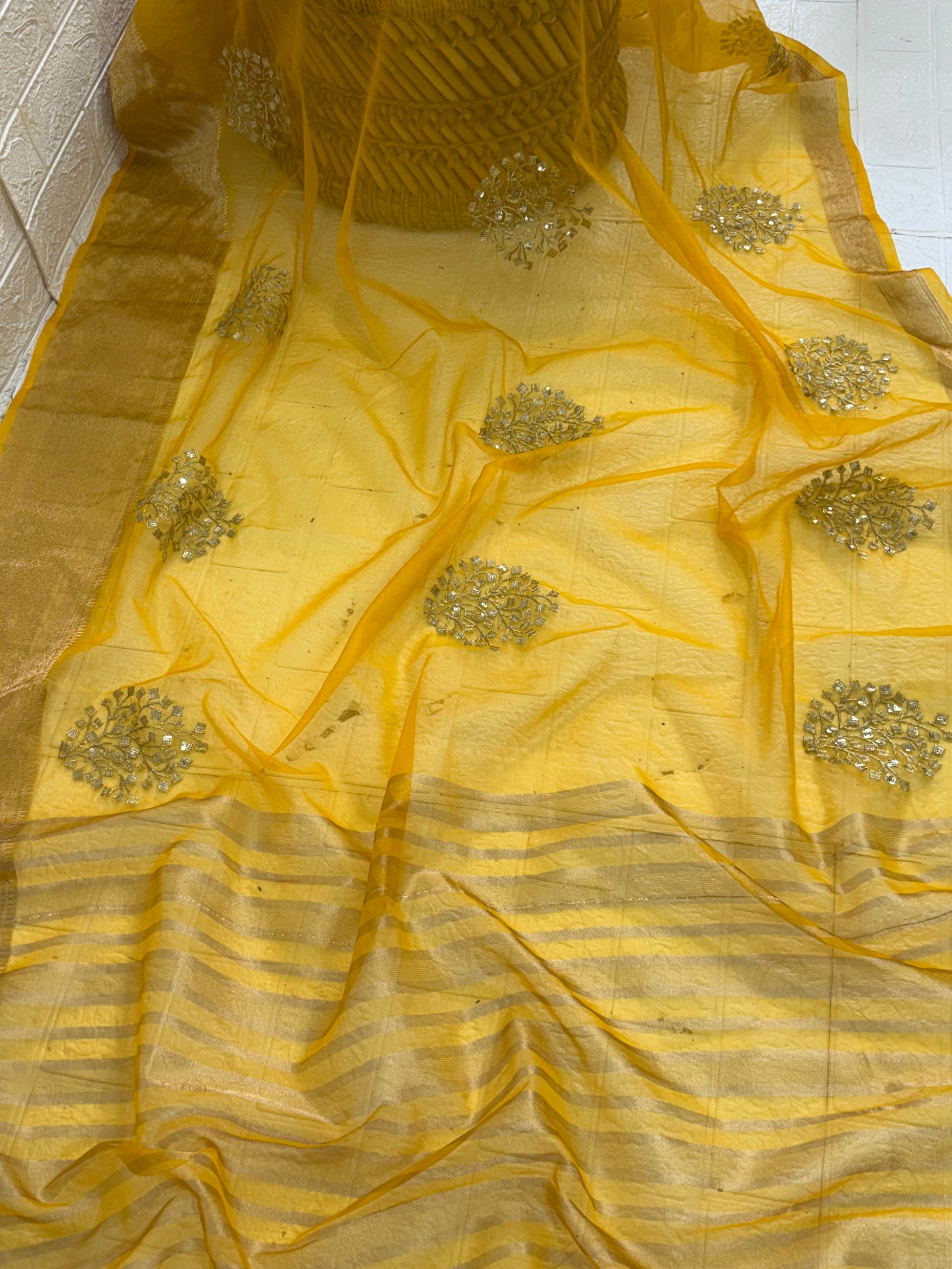 This is likely an Organza or fabric saree. The sheer, light-weight look suggests a fine fabric like Organza, or a similar silk blend, adorned with matching blouse golden zari lines on the pallu and intricate golden sequin or bead floral embroidery