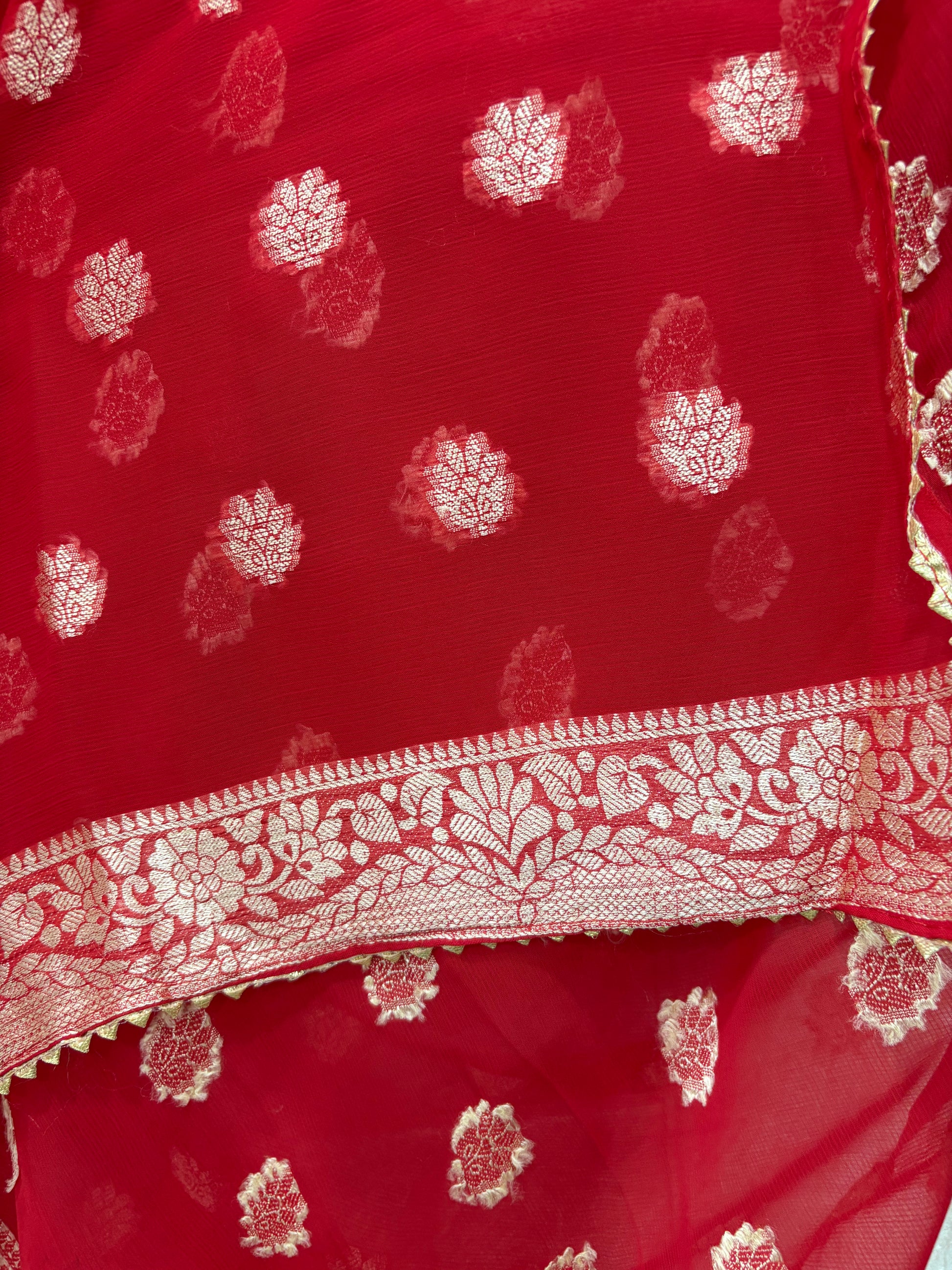 This fabric is a red Georgette with all-over gold zari floral buti embroidery and a rich woven border, paired with a matching plain blouse perfect for festival